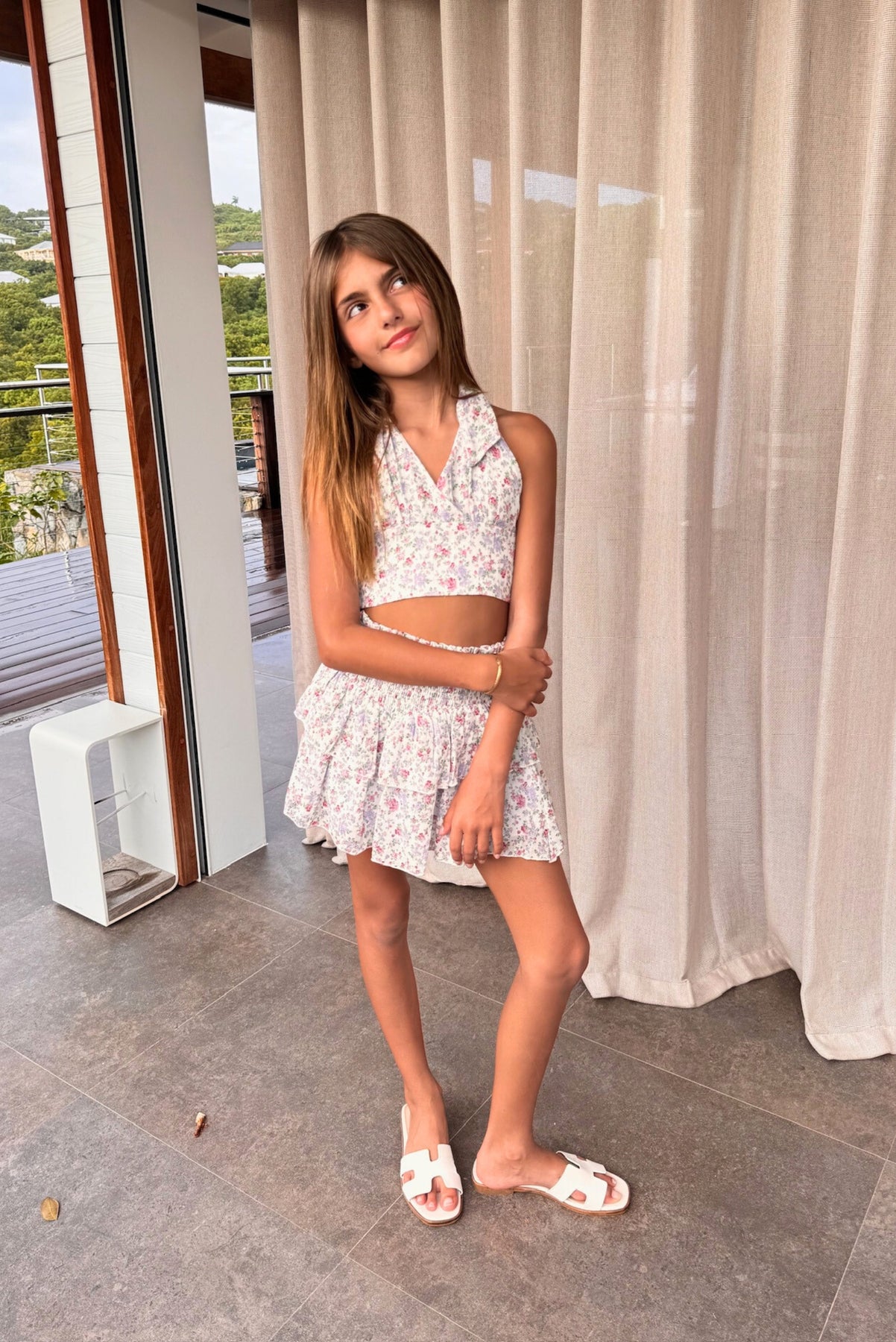 a young girl stands indoors in a stylish white floral halter top and matching skirt. she's wearing white sandals and is looking up. the background features curtains, a window with a view of a balcony and a deck beyond. the overall setting is a wellappointed home with light and airy decor.  the scene is bright and pleasant.