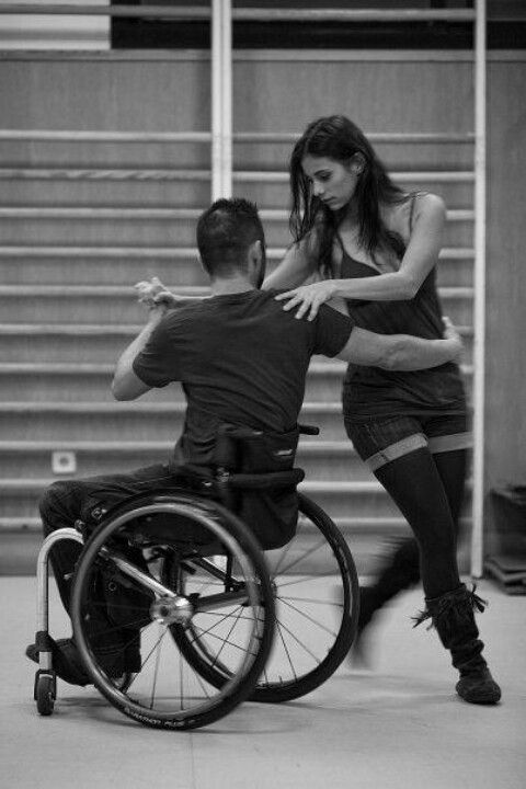 a black and white image of a man and a woman dancing. the man is in a wheelchair and the woman is holding onto him as they sway. they are both smiling and appear to be in love.  the background is a blurry wall with lines going vertically, like the beams in an old gym or dance studio.