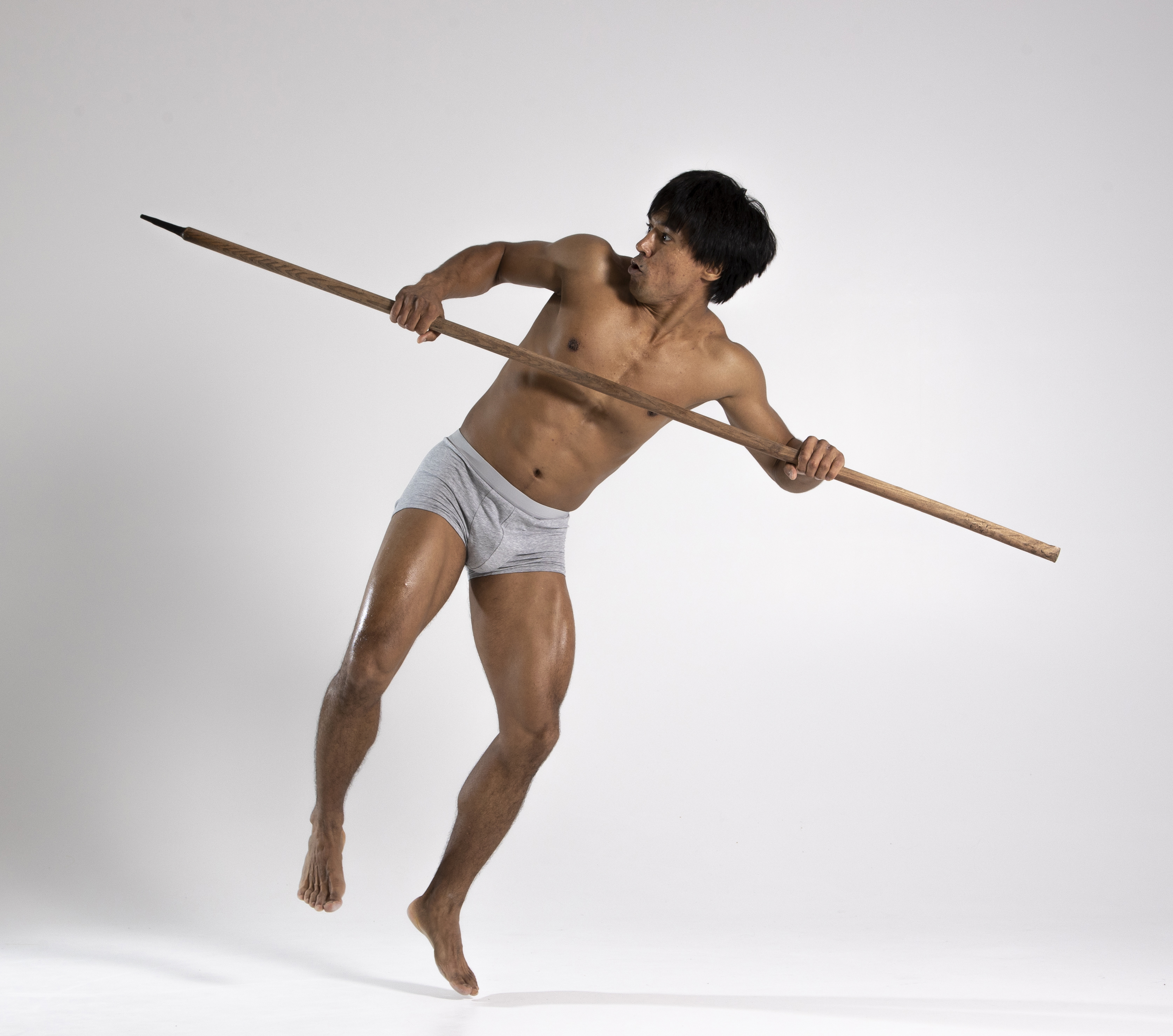 a muscular, shirtless man in gray underwear is posed in a dynamic action stance against a light gray background. he holds a long wooden spear horizontally, gripping it with both hands. his feet are bare, and his body is angled as if in motion or engaged in a defensive maneuver. the scene appears to be a studio shot emphasizing his physique and the weapon he wields.
