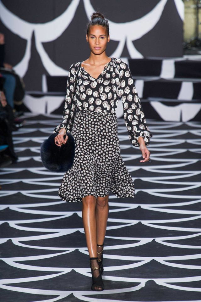 a fashion model walks down a runway wearing a black and white floral print dress. the dress has a skirt and blouse, and the model also has on a fur handbag, black ankle boots and a bun. the runway has a black and white wavy design.