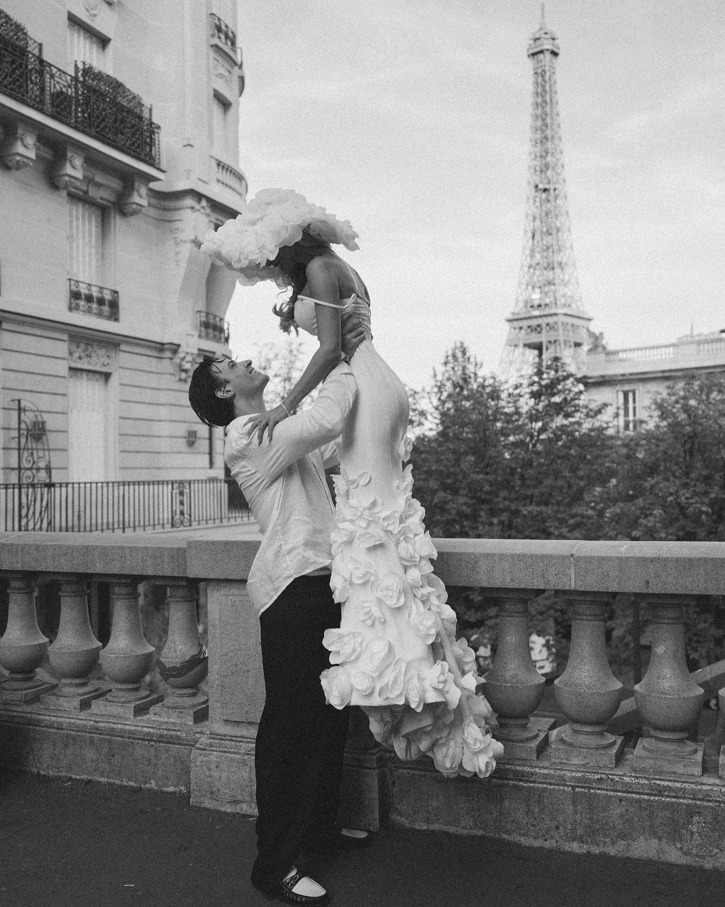 a couple in paris. the man is holding the woman in his arms and is giving her a kiss. they are standing in front of the eiffel tower. the woman is wearing a white dress and a hat.  the man is wearing a white shirt and black pants.  the image is in black and white.