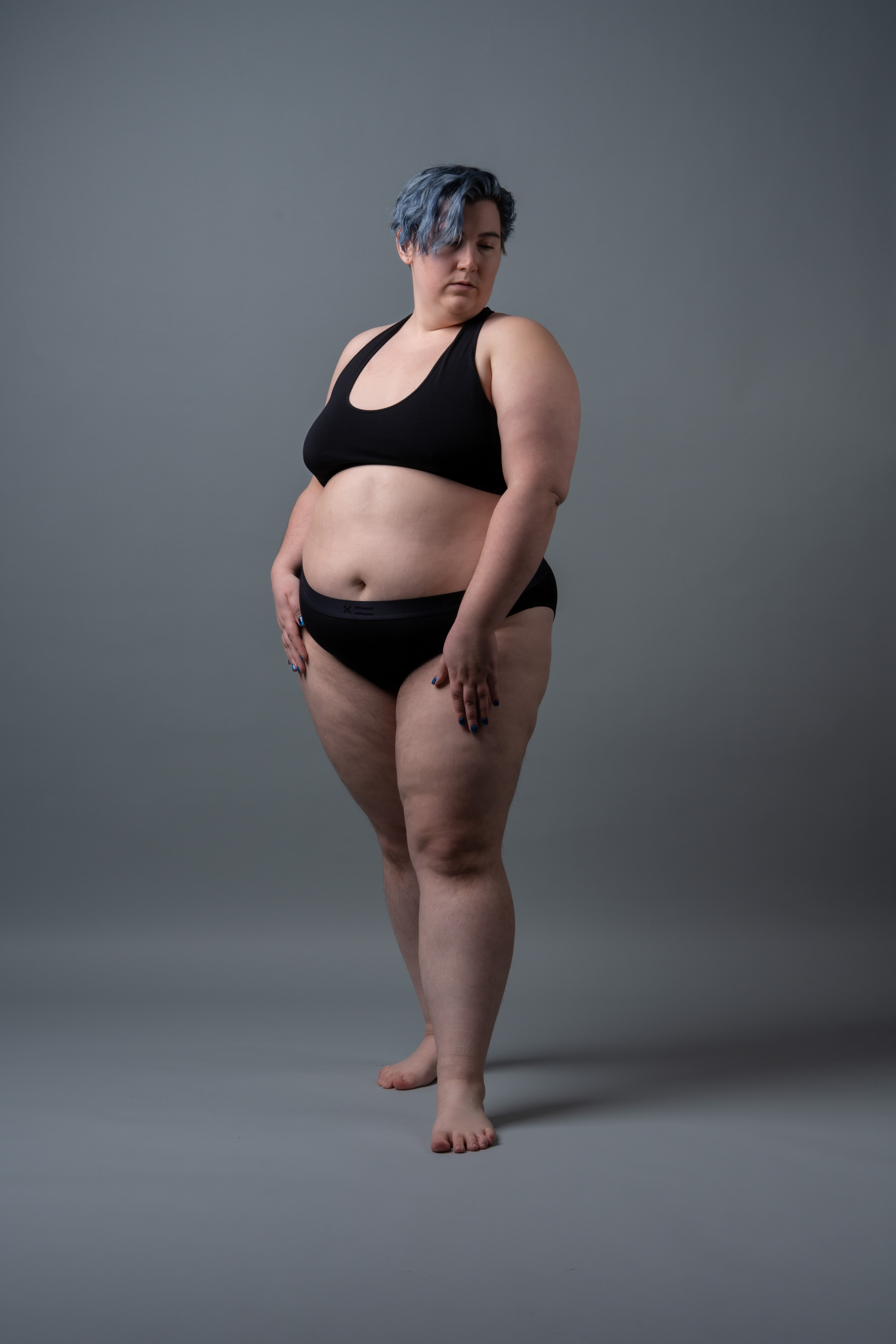 a full body studio shot of a plus size woman wearing a black sports bra and black underwear, standing barefoot against a gray background. she has short blue hair and is looking down.