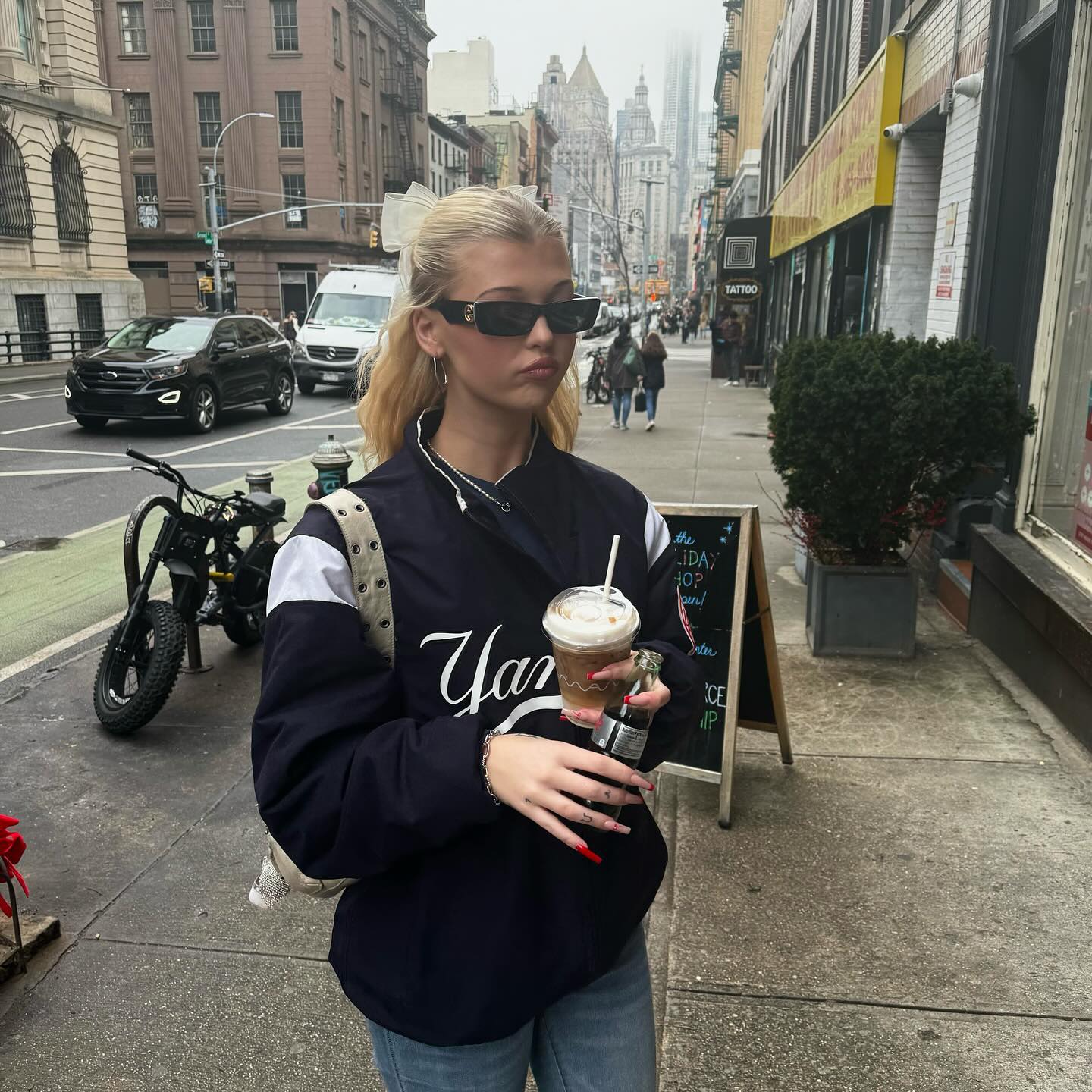 a woman with blonde hair is standing on a sidewalk in a city, wearing sunglasses and a yankee jacket, holding a cup of coffee and a bottle. there are buildings, a tree, a motorcycle, a street, and a black car in the background.