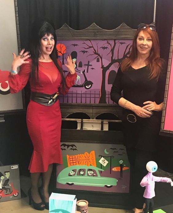 two women are standing in front of a large art piece. the woman on the left is dressed in a red dress with a black belt. she is smiling and her hands are raised. the woman on the right is dressed in a black shirt and black pants. she is smiling but with her hands clasped in front of her.  there is a painting of a spooky scene with a car driving in the middle. it appears to be a halloween themed art exhibit.