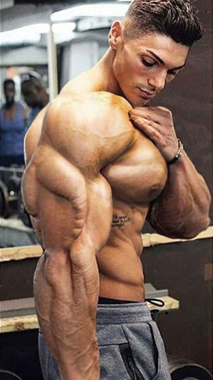 the image shows a muscular man, possibly a bodybuilder, in a gym setting. he is shirtless, showcasing his highly developed muscles, including his biceps, triceps, shoulders, chest, and abs. his physique is extremely welldefined, with visible veins. he has short, dark hair and a tattoo on his side. he's wearing grey sweatpants and appears to be posing, possibly for a photograph. the background is blurry but shows a gym environment with mirrors and other equipment. the focus is on the man's impressive muscular build. the image is a closeup portrait, highlighting the detail of his muscles and skin.