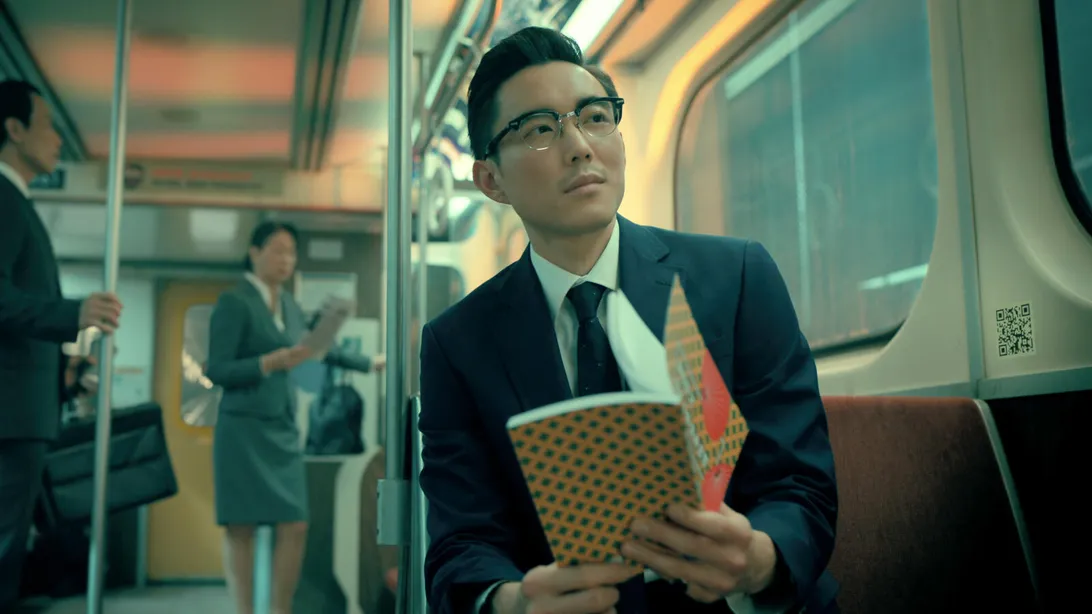 a man in a suit and glasses sits on a bustling subway train. he holds a patterned notebook and a brightly colored object, possibly a book or folder, in his hands. he's looking off to the side, appearing lost in thought. the lighting is bright and artificial, typical of a subway car. in the background, other passengers stand, including a woman in a suit. the overall atmosphere suggests a busy urban commute.