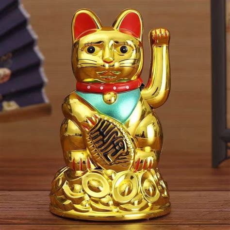 closeup view of a golden manekineko, or beckoning cat, figurine. the cat is seated with one paw raised, a traditional pose associated with good luck and prosperity. it's adorned with red and teal accents, and rests on a base designed to look like a pile of gold coins. the figurine appears to be made of a shiny, goldcolored material, possibly plastic or resin.  the background is blurred, showing hints of a wooden surface and other decorative elements.
