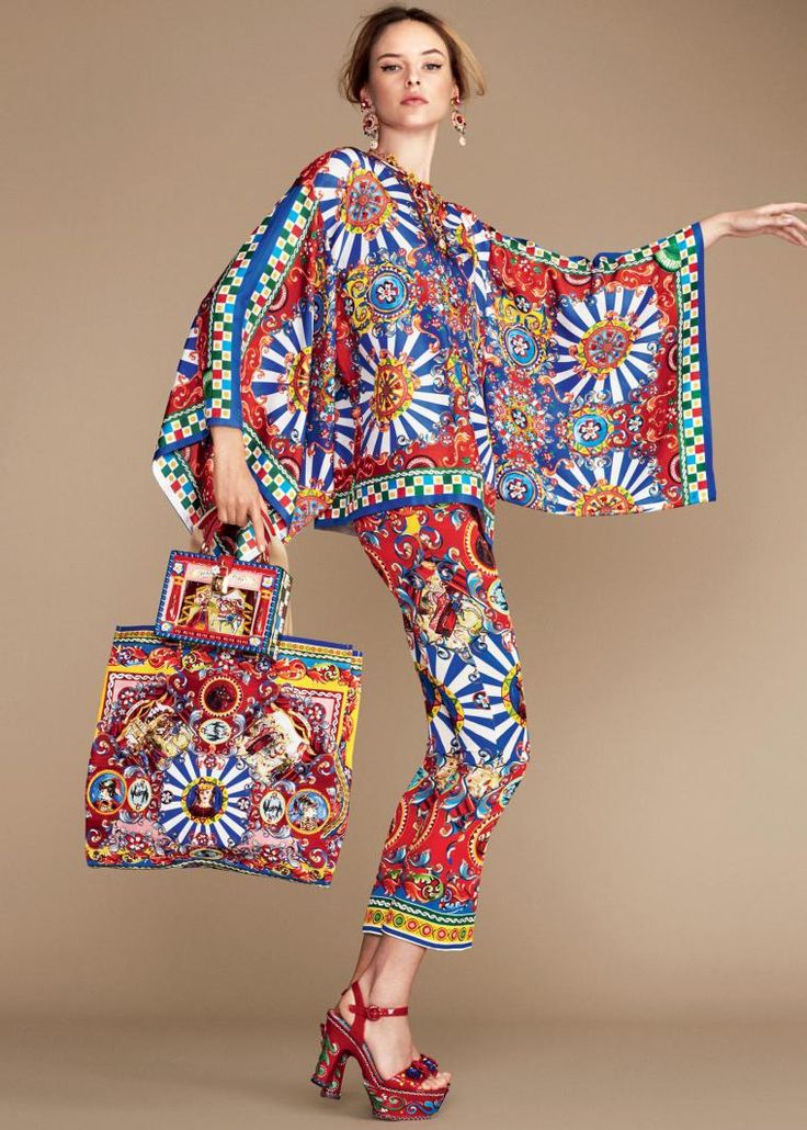the image showcases a fashion model wearing a striking dolce & gabbana outfit. the ensemble consists of vibrant, wideleg trousers and a matching loosefitting,  ponchostyle top, both adorned with a rich, multicolored baroqueinspired print featuring florals and swirling patterns. the model also carries a large tote bag with a coordinating print. she is wearing red platform heels and delicate earrings, completing a stylish and luxurious look. the background is a simple, neutral beige, allowing the clothing to take center stage. the overall style is elegant, expressive, and characteristic of dolce & gabbana’s signature aesthetic.