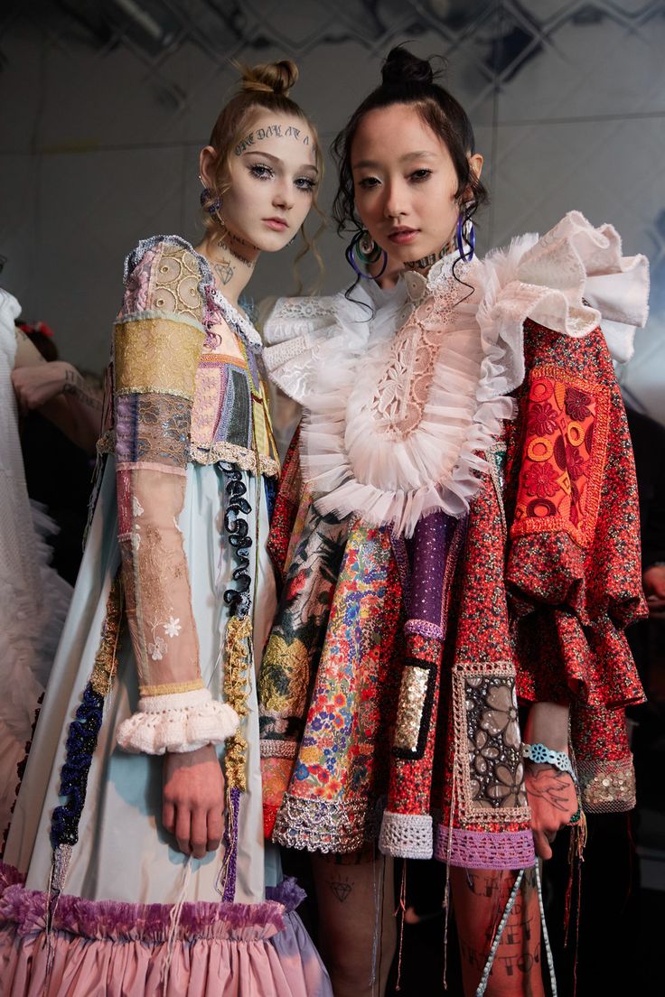 the image showcases two fashion models backstage at a fashion show, likely a runway presentation of avantgarde or haute couture clothing.  both models are adorned in extremely detailed and elaborate dresses.  the dresses are a patchwork of fabrics, featuring embroidery, lace, and various other embellishments.  the styles are highly eclectic and unique, blending seemingly disparate elements together. the models' makeup and hairstyles add to the overall artistic and slightly rebellious feel. one model's face is partially adorned with small tattoos.  the overall impression is one of highfashion artistry and individualistic style.