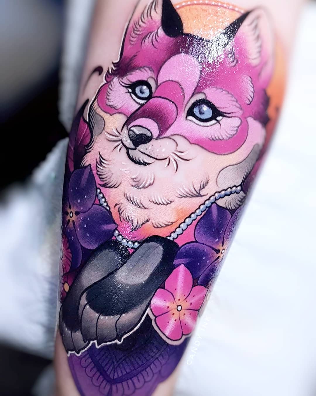 a tattoo of a pink fox with blue eyes and black paws. the fox has a necklace of white pearls and there are purple flowers around the fox's neck and paws. the tattoo has a purple background.