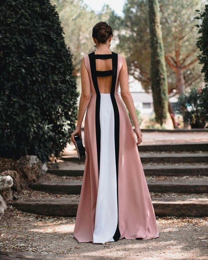 a woman stands on a set of wooden steps in an outdoor setting, wearing a striking backless gown. the dress features a colorblocked design with panels of pink, white, and black. her hair is styled in an updo, and she is holding a clutch in her left hand. the background includes trees and greenery, suggesting a garden or park.