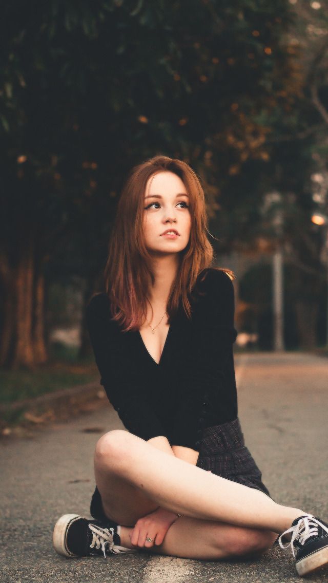 a young woman with long, copperbrown hair sits on a sidewalk with her legs crossed. she is wearing a black longsleeved shirt and a black and white skirt. her black and white vans sneakers are untied. she is looking off to the right, and her expression is pensive. the background is a street lined with trees, and the lighting is soft and warm, suggesting it is evening.
