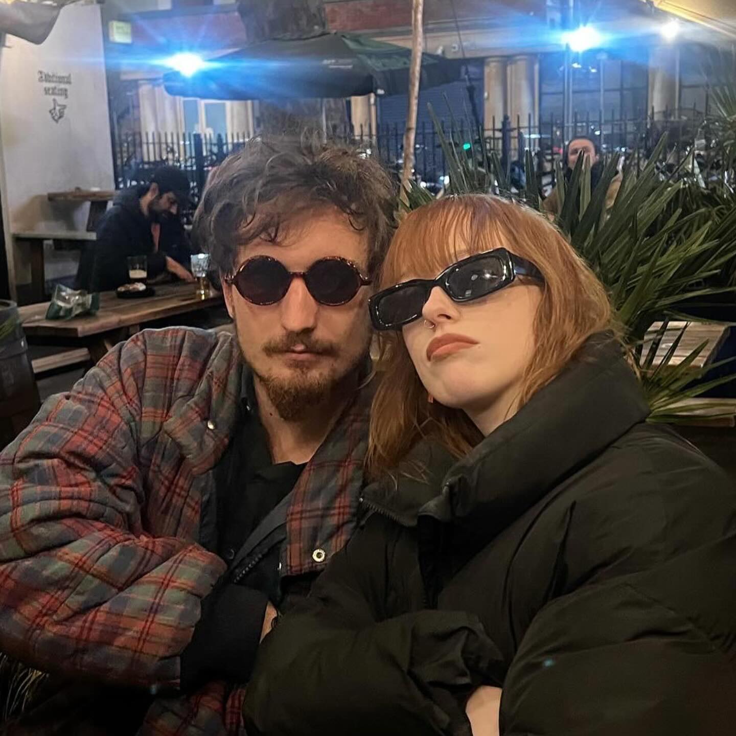 a closeup shot of a young couple sitting outside at a pub at night. the man is wearing a red and green plaid shirt and brown sunglasses, while the woman is wearing a black puffer jacket and black sunglasses. they are sitting close together, and the woman's head is resting on the man's shoulder. the background is slightly blurred but shows a city street with other people and buildings visible. the overall mood of the image is romantic and casual.