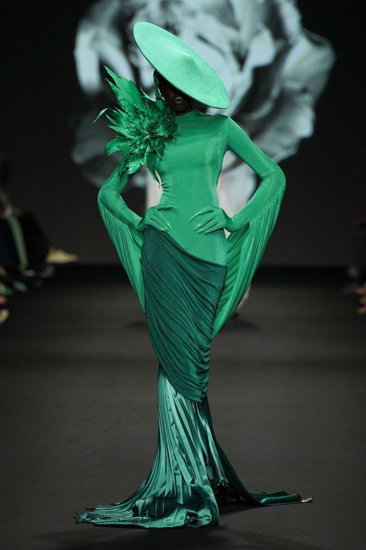 a model in a green dress and a green hat walks the runway. the dress is long and flowing, with a pleated skirt that is cascading towards the floor. she is posing with her hands on her hips and her body is slightly arched. the background is a blurred image of a white flower.
