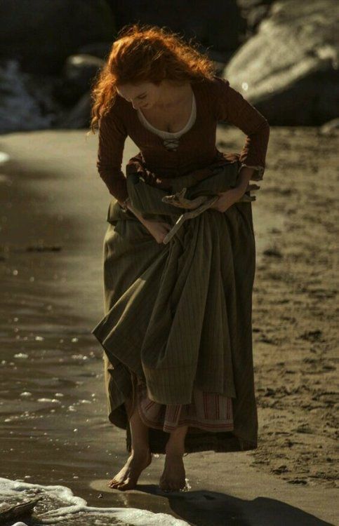 a young woman with long, red hair stands on a beach at the water's edge. she is wearing a long, brown dress and is looking down at the sand. the woman is holding a piece of driftwood in her hands. the lighting is warm and natural, and the overall feel of the image is one of peace and serenity.