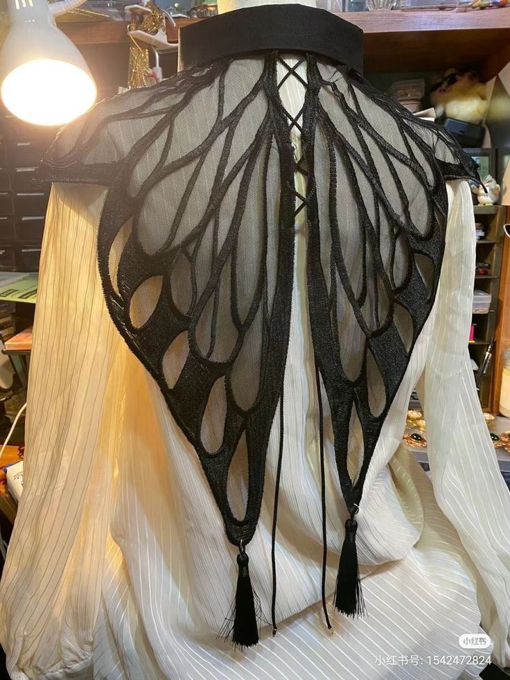 a white blouse with a black butterfly wingshaped collar. the collar is made of a sheer material with black trim and has tassels hanging from the bottom. the collar is laceup in the back. the blouse has long sleeves and a slight sheen. a lamp provides light from the upper left.