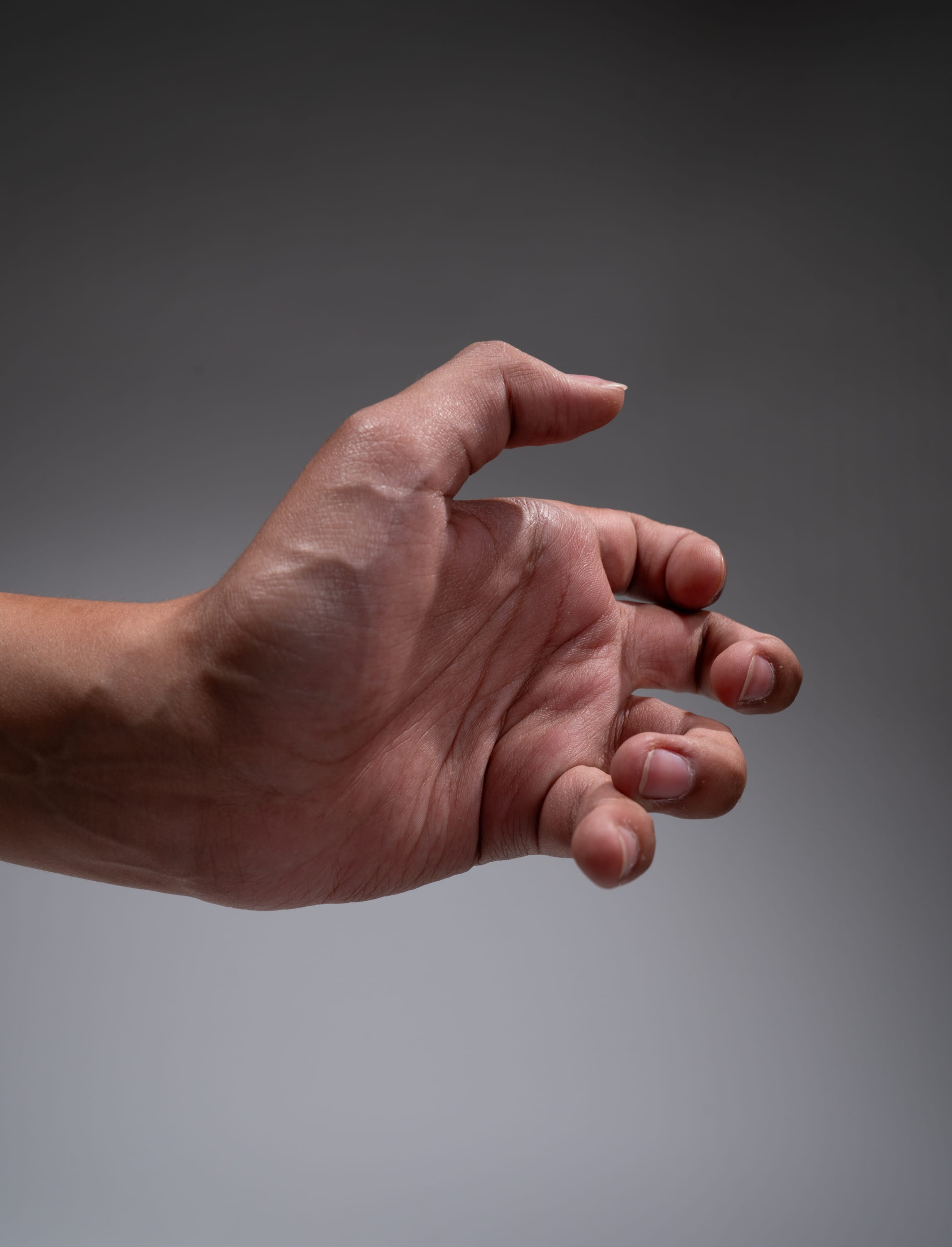 a closeup shot of a person's right hand extended forward, as if reaching or grabbing for something. the hand is slightly cupped, with the fingers curled inward. the skin of the hand is tanned, with prominent veins and wrinkles, suggesting maturity and perhaps manual labor. the lighting highlights the texture and form of the hand, casting subtle shadows that enhance its dimensionality. the gray background isolates the hand, drawing attention to its gesture and details.
