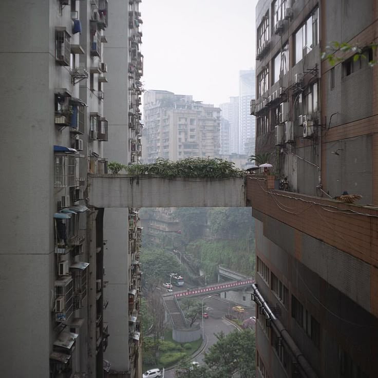 the image shows a narrow alleyway between two tall, grey apartment buildings in a dense urban setting.  a concrete bridge or walkway connects the two buildings at a higher level, creating a unique architectural feature. the view beyond the buildings shows a busy street with cars and some green vegetation, all shrouded in a hazy, misty atmosphere.  the overall mood is one of urban density and a somewhat claustrophobic feel, highlighting the close proximity of the buildings and the restricted perspective.  the scene suggests a location in a rapidly developing city, perhaps in asia.