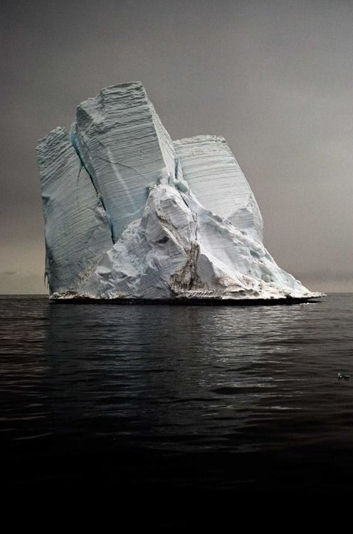 the image shows a large iceberg floating in the dark ocean under a grey sky. the iceberg is massive, with vertical and horizontal grooves along the surface.