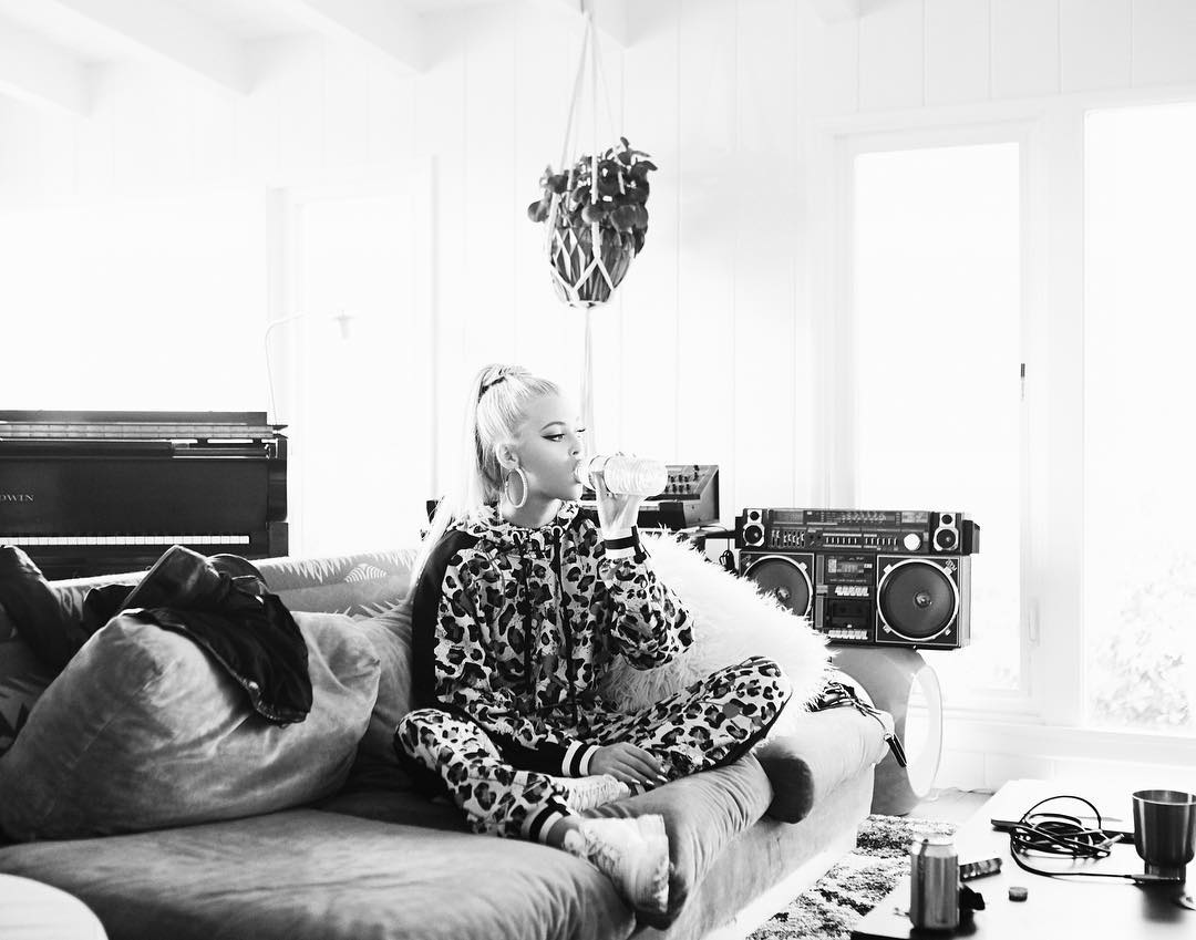 a black and white photograph features a woman sitting crosslegged on a couch in a living room. she is wearing a leopard print tracksuit and large hoop earrings, and she is drinking from a water bottle. to her right is a large boombox sitting on top of a drum, and behind her to the left is a piano. an indoor plant hangs from the ceiling above her. the living room is brightly lit, with windows in the background. the image has a stylish and relaxed vibe.