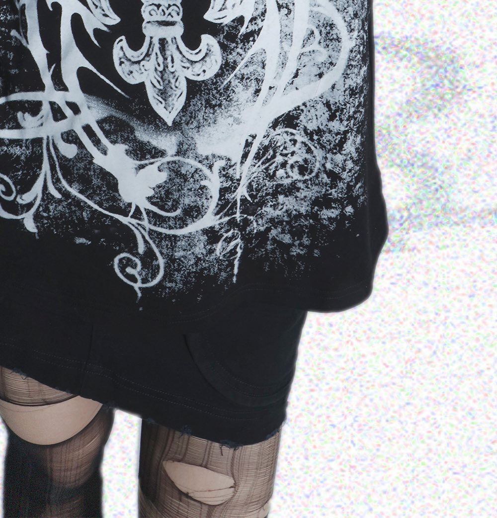 the image shows a closeup of a person wearing a black, oversized tshirt dress featuring a large, distressed white print of a fleurdelis and ornate floral designs. the shirt has a slightly worn look, and the person is also wearing fishnet tights with visible rips and tears. the background is a slightly blurry, lightcolored texture. the overall style is gothic, grunge, or punkinspired.