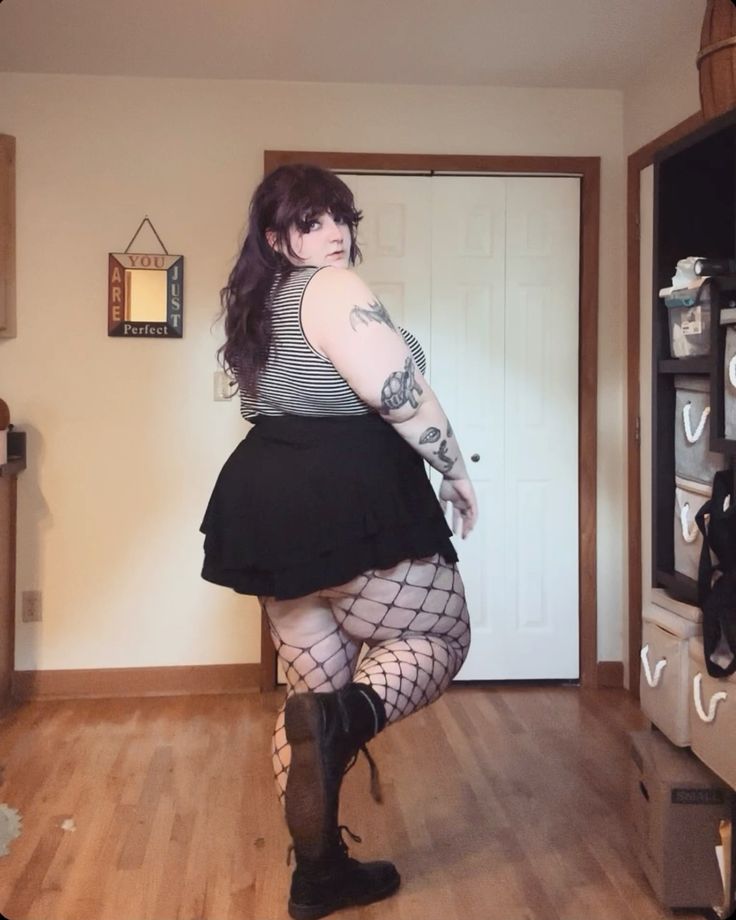 a woman with purpletinted hair is standing in a room. she is wearing a black and white striped tank top, a black skirt, fishnet stockings, and black boots. she has tattoos on her arm.