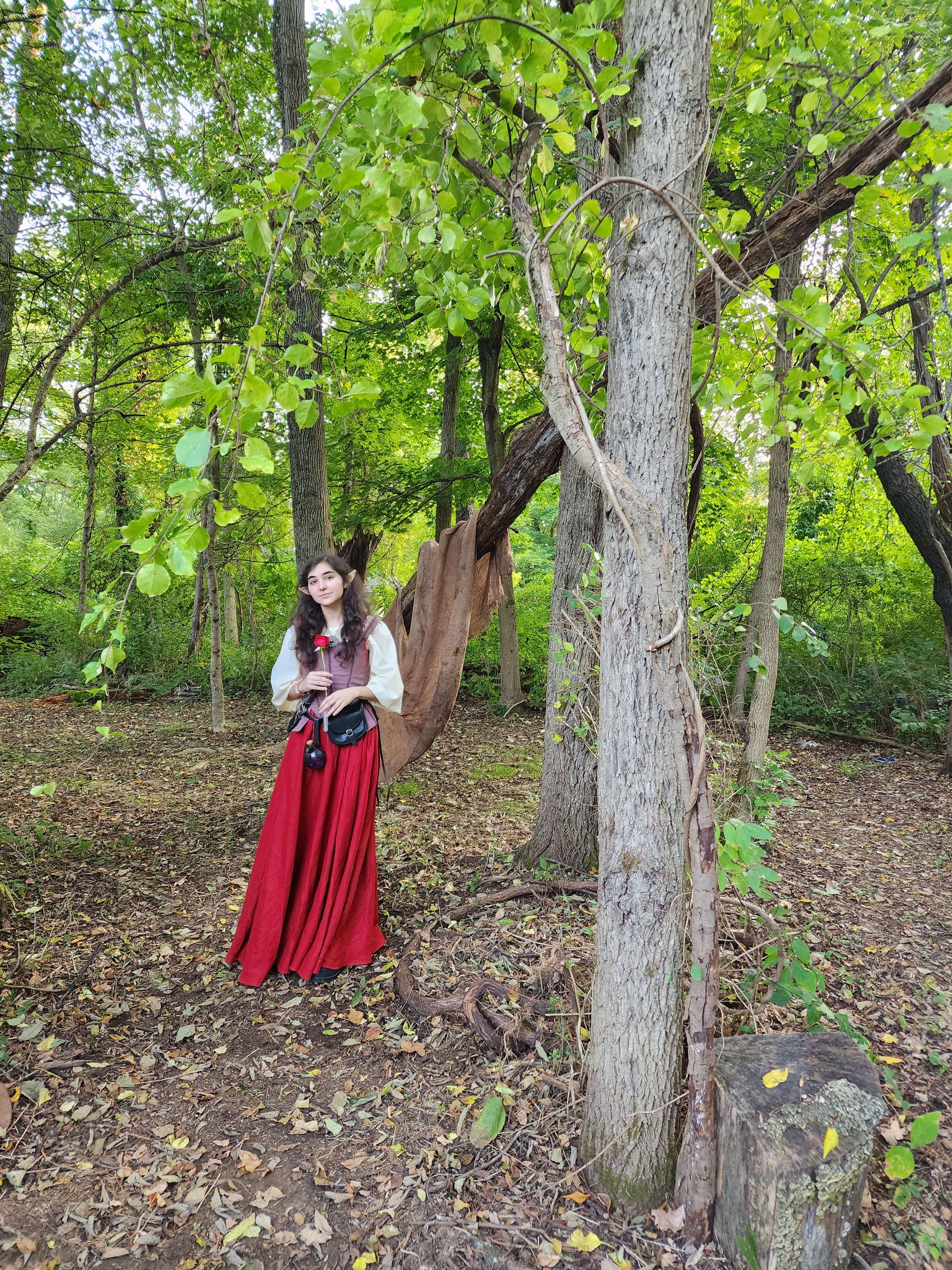 a young woman with long brown hair, wearing a long red medievalstyle dress and a creamcolored bodice, stands in a forest. she holds a single red rose in her hand. the forest is lush with green leaves and the ground is covered with fallen leaves. a large tree trunk is prominent in the image, with a piece of brown fabric draped over a branch.  she appears to be posing for a picture.  the overall atmosphere is whimsical and slightly magical.