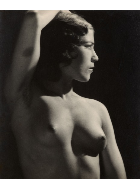 a black and white photograph of a nude woman, viewed from the side, her arm raised above her head. the image features strong contrasts of light and shadow, highlighting the curves of her body. her hairstyle suggests the 1920s or 1930s. the background is dark and undefined, focusing attention on the subject. the overall style is reminiscent of classic artistic nude photography from the era.