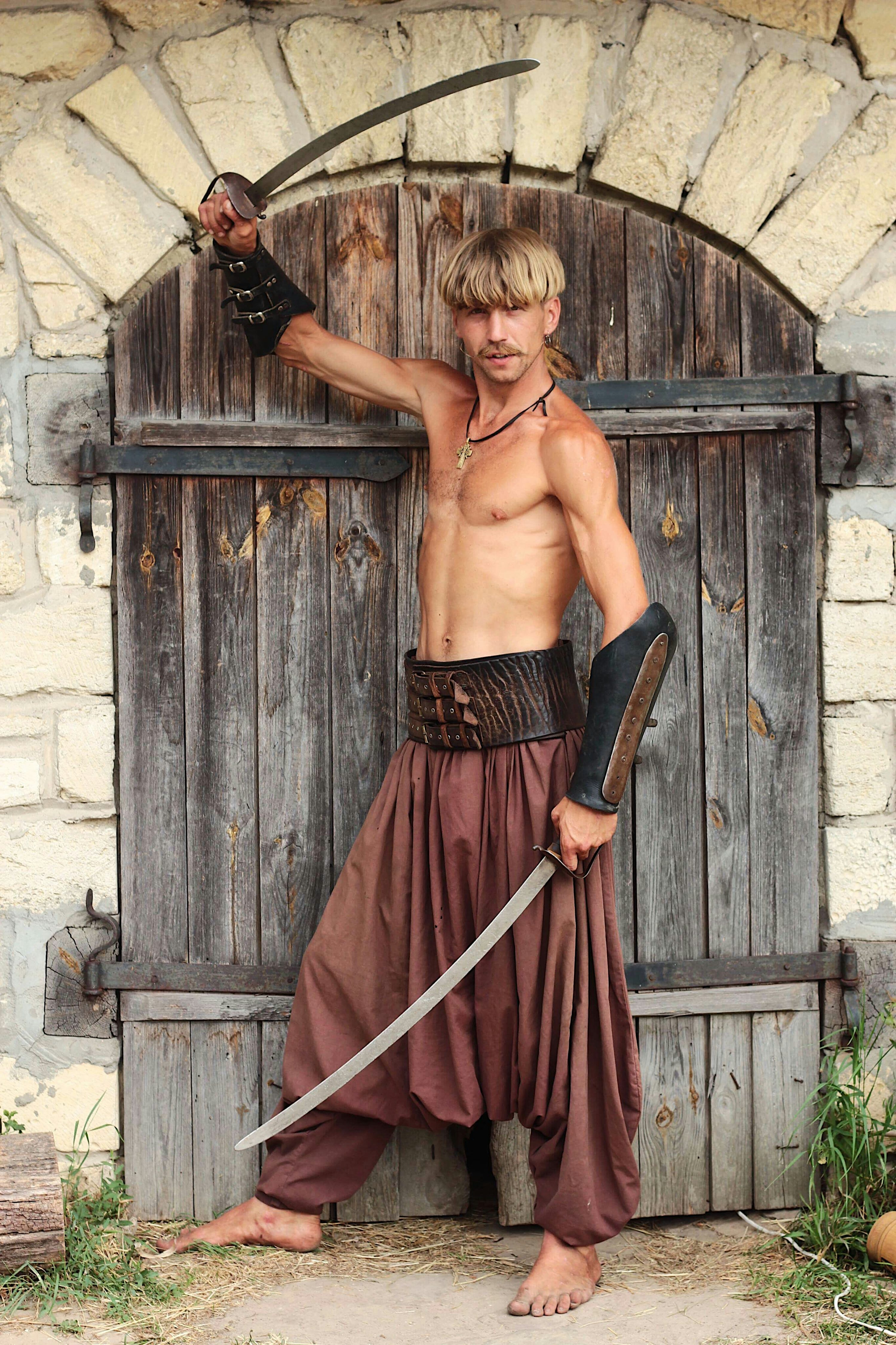a shirtless, muscular man, possibly portraying a cossack warrior, stands barefoot before a rustic wooden door set within a stone wall. he is holding a curved saber (shashka) and is wearing traditional clothing including brown harem pants, a leather belt, and leather arm guards.  the image is a fullbody portrait with the man posing confidently. the scene is suggestive of a historical or traditional setting.