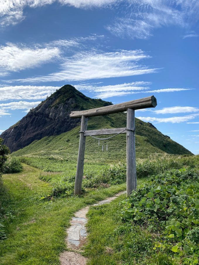 a scenic landscape featuring a mountain backdrop and a traditional wooden torii gate in the foreground. a stone path leads through lush greenery towards the gate. the sky above is blue with scattered clouds.