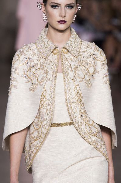 loseup view of a female model showcasing a luxurious, intricately embroidered cape and dress ensemble. the cape is offwhite or ivory with elaborate gold embroidery creating detailed floral and geometric patterns.  the dress underneath is a matching solid color, appearing to be made of a fine woven material. the model wears delicate gold earrings and a simple gold belt that is barely visible under the cape.  the background is blurred, suggesting a fashion runway setting. the overall impression is one of highend fashion and exquisite craftsmanship.