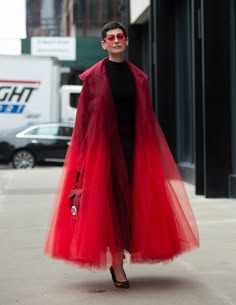 a woman wearing a long red dress with tulle skirt is walking down the street in a city environment. she is wearing sunglasses and has a confident look on her face. she looks chic and stylish.