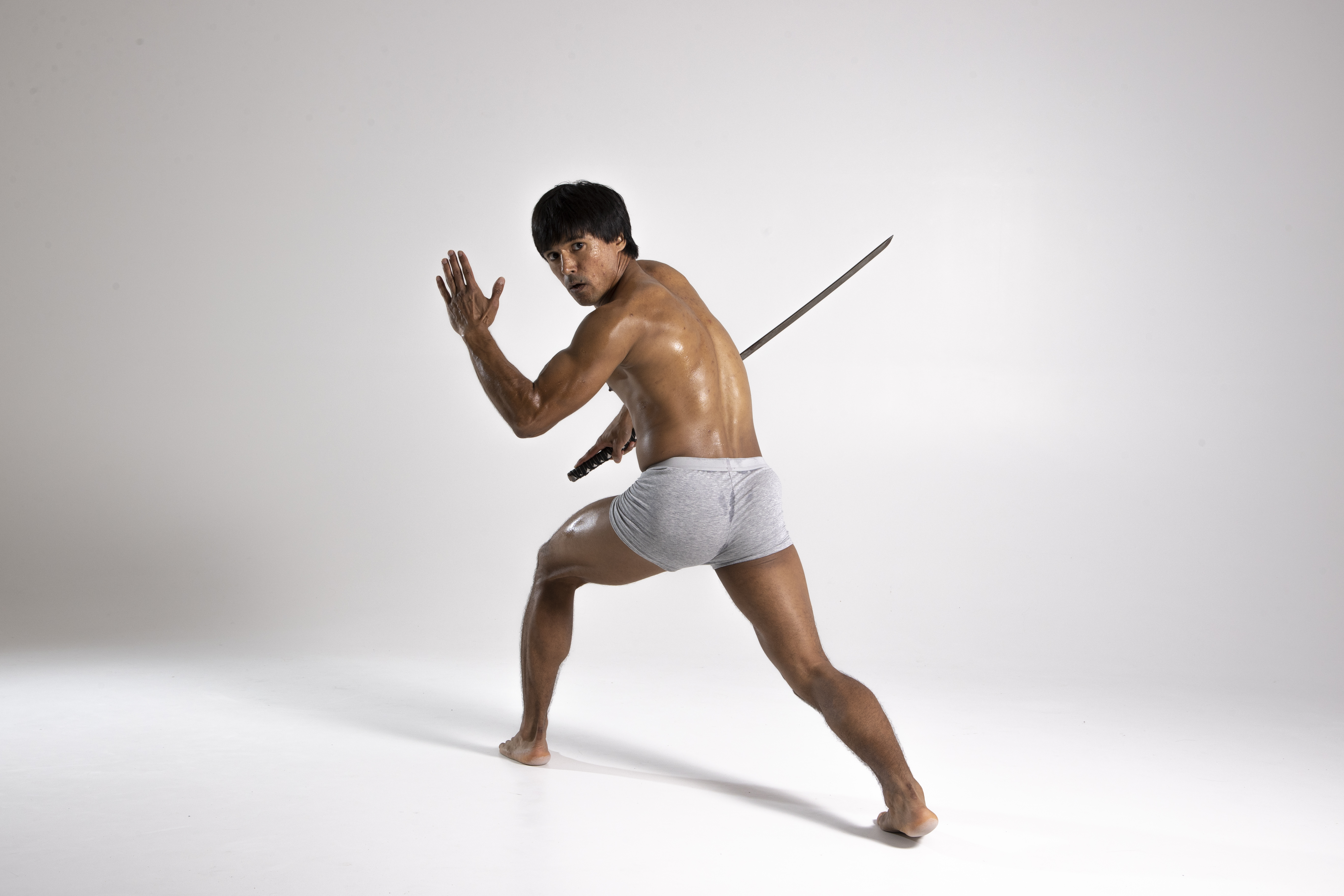 a muscular, shirtless man wearing a loincloth is posing with a katana sword. he is barefoot and looking over his shoulder with a determined expression. the background is a plain white studio setting. he is in a defensive martial arts stance.