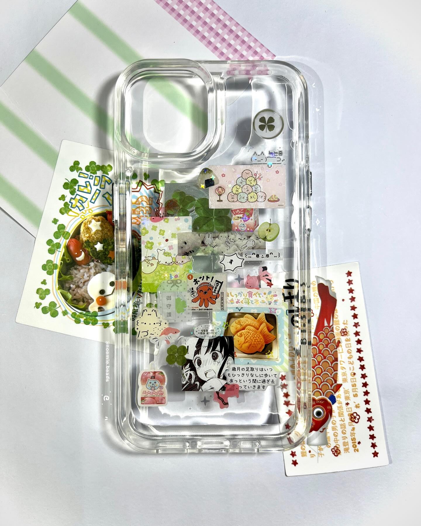 a clear phone case decorated with a variety of japanesethemed stickers. the stickers include images of food, anime characters, and traditional symbols like koi fish and clovers. the phone case is sitting on a surface with additional cards and decorative paper.