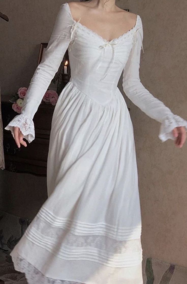 a woman wearing a long white dress stands in a room. the dress is made of a lightweight fabric and has long sleeves, a sweetheart neckline, and a flowing skirt. the dress is adorned with lace trim. the woman is posing with her arms outstretched. the room is decorated in a vintage style. the lighting is soft and natural.