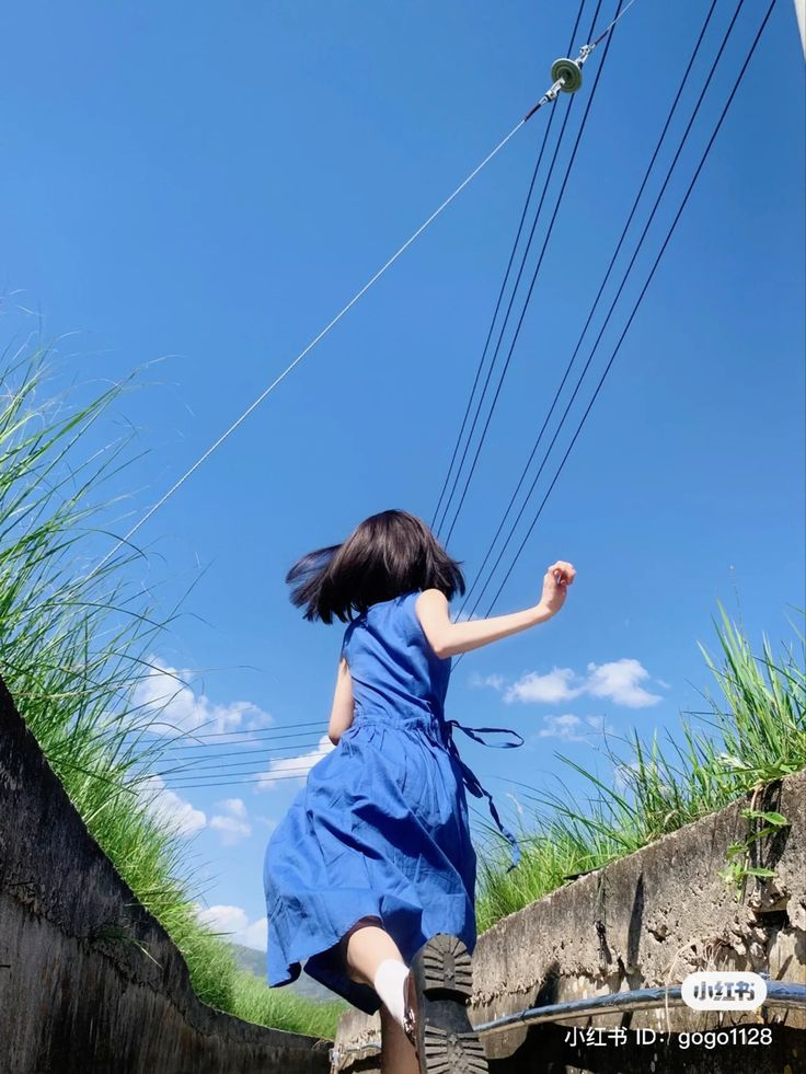 a woman wearing a blue dress and shoes runs outdoors during the day with a blue sky overhead. power lines are visible in the sky. the woman is surrounded by grass and concrete structures.