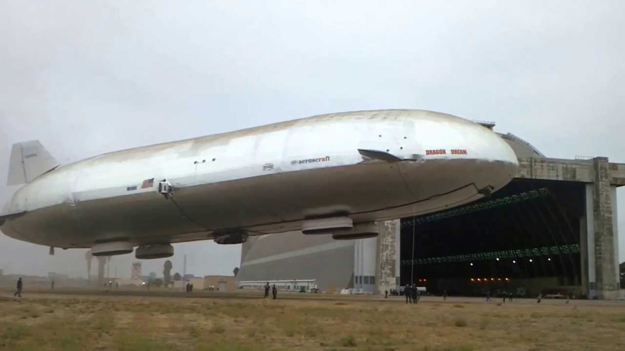 a large airship, or blimp, is parked outside a hangar. the airship is white with red lettering. the hangar is grey and made of concrete. there are some people standing outside the hangar. the sky is cloudy.