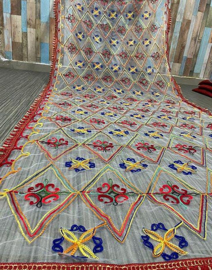 the image shows a sheer dupatta, likely a traditional indian or pakistani scarf, laid out flat. it features a geometric diamond pattern with intricate embroidery in various colors including red, blue, yellow, and green. each diamond contains either a floral motif or a more abstract design. the dupatta is finished with a red tassel trim along its edges. the fabric appears to be lightweight and delicate.