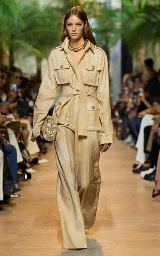 a blonde model walks down a runway wearing a long tan jacket and wideleg pants. the outfit is styled with a gold necklace and a tan clutch. the model is posing for the camera with a confident expression.