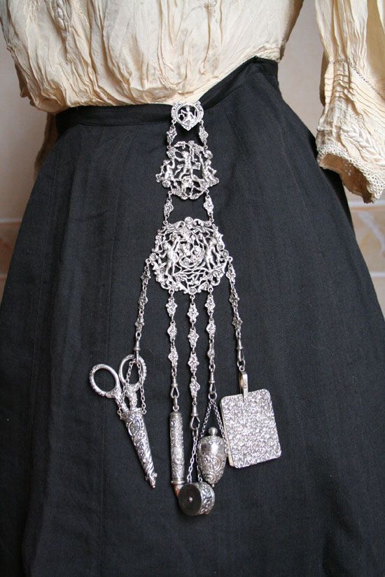 the image shows a woman's torso, wearing a lightcolored, ruffled blouse and a dark, highwaisted skirt. a silver chatelaine is attached to the waistband of the skirt. the chatelaine consists of a series of decorative filigree links and chains that suspend various items, including a pair of scissors in a sheath, a needle case, a small perfume bottle, and a small square notebook. the chatelaine is made of silver and features intricate metalwork.