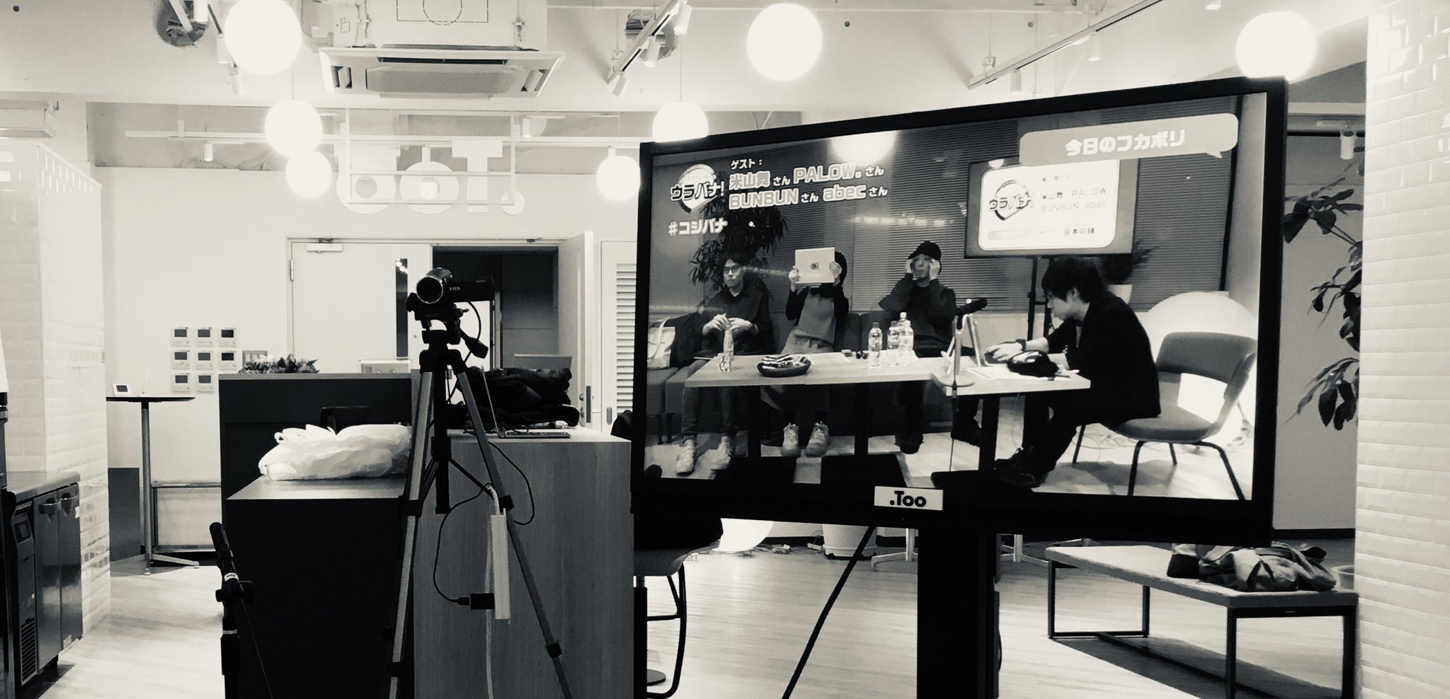 the image shows a black and white photograph of a television studio set up for a talk show or broadcast.  a large flatscreen tv displays four individuals seated around a table, engaging in a discussion.  the environment appears to be a modern office space, with overhead lighting illuminating the set.  a camera on a tripod is positioned to capture the broadcast from an eyelevel perspective.