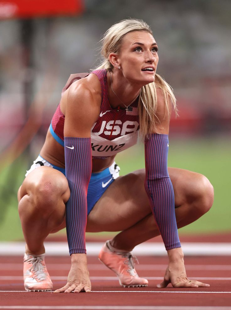 a female athlete wearing a usa uniform is crouched down on a track and field track, ready to start a race. she is looking straight ahead with a focused expression.