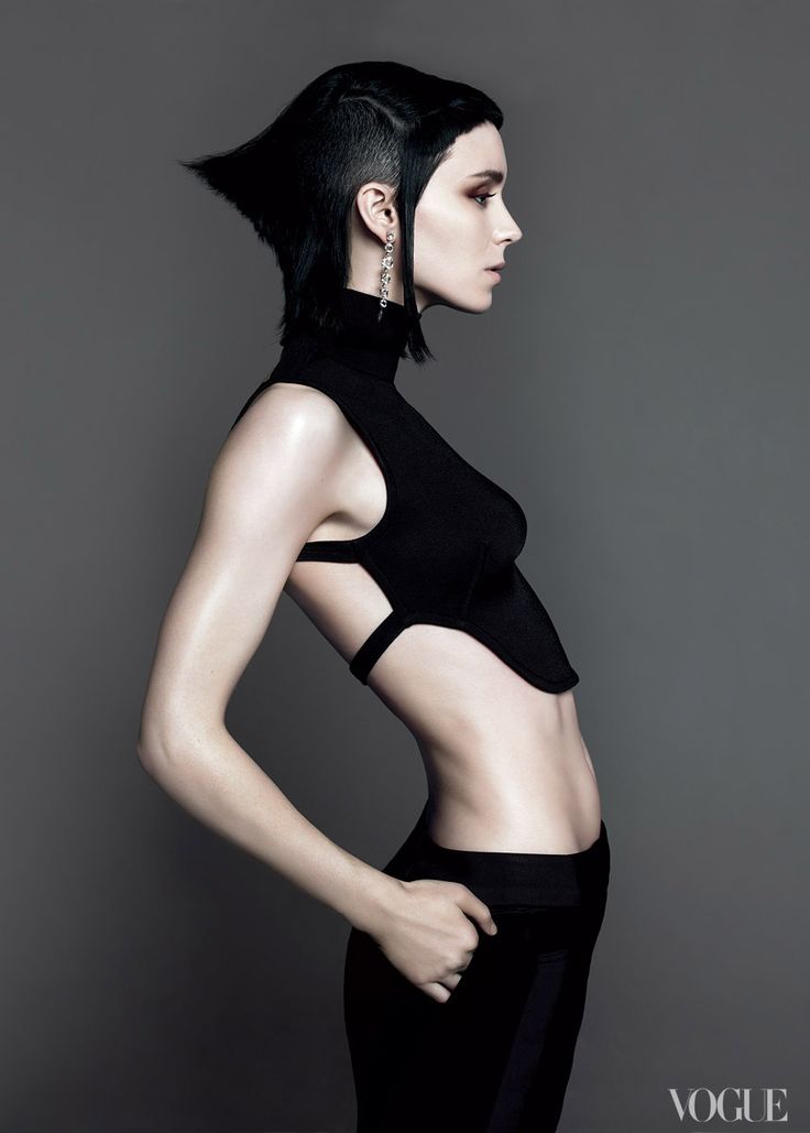 a striking black and white fashion photograph features a young, thin woman with a unique, edgy haircut and minimal makeup. she's wearing a black, sleeveless top with cutouts on the sides, paired with black pants. the model is posed with her body slightly turned to the side, offering a captivating profile view against a plain grey background. the image exudes a sense of high fashion and contemporary style.