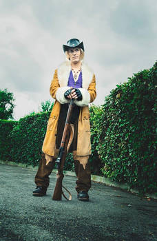 a fulllength shot of a person in a cowboy/cowgirl costume, possibly cosplay, standing on a paved path beside a green hedge. they are wearing a brown furtrimmed coat, a cowboy hat, and brown boots. the person is holding a long rifle and has a serious expression on their face. the background is an overcast sky and the overall setting appears rural. the image has a vintage or retro aesthetic.