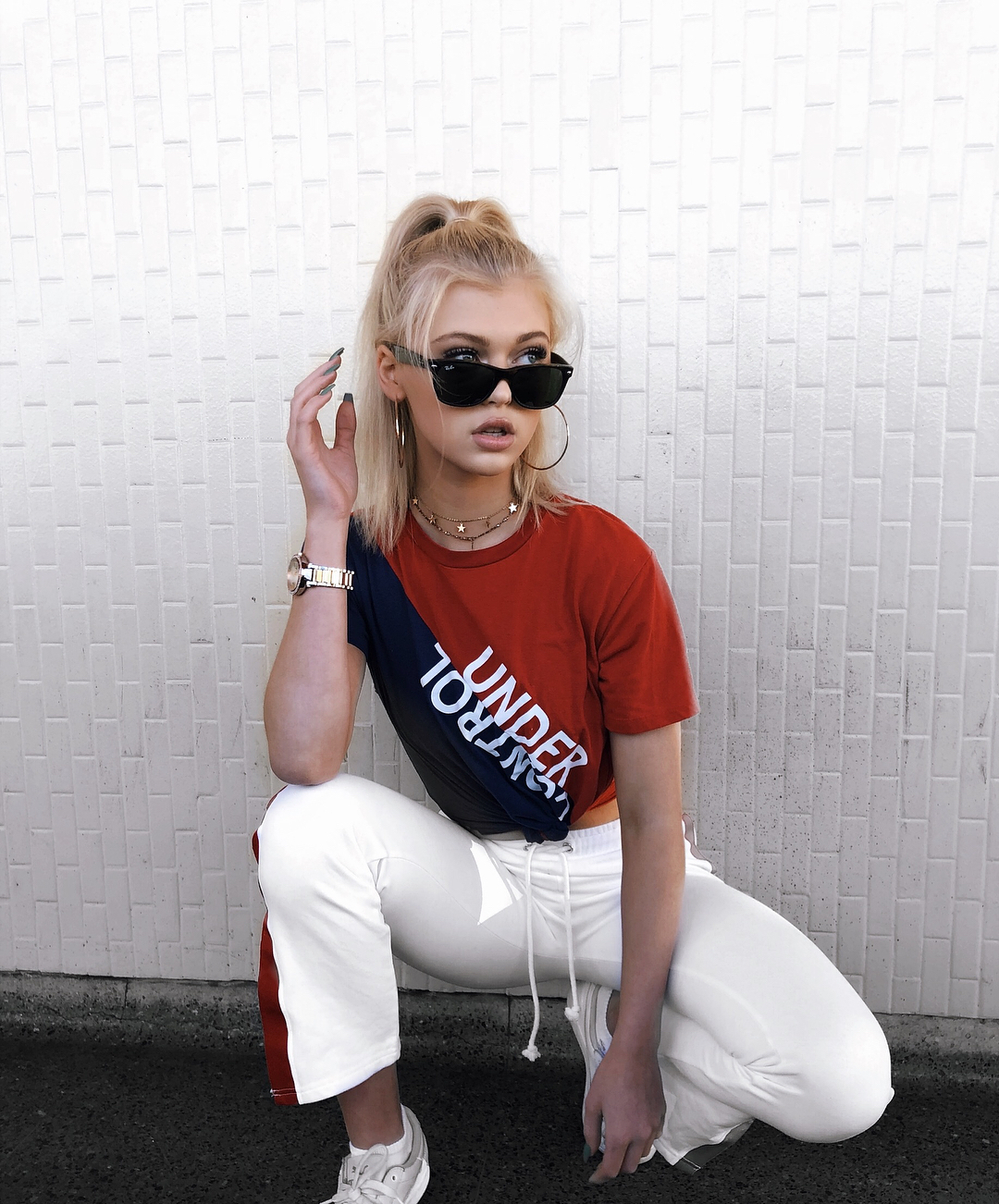 a young woman with blonde hair in a high ponytail, wearing sunglasses, a red and navy tshirt with the words "under control", white sweatpants with a red stripe down the side, and white sneakers, is crouching against a white brick wall. she is wearing jewelry including a watch, necklaces, and hoop earrings. she is looking to the side with her hand near her face.