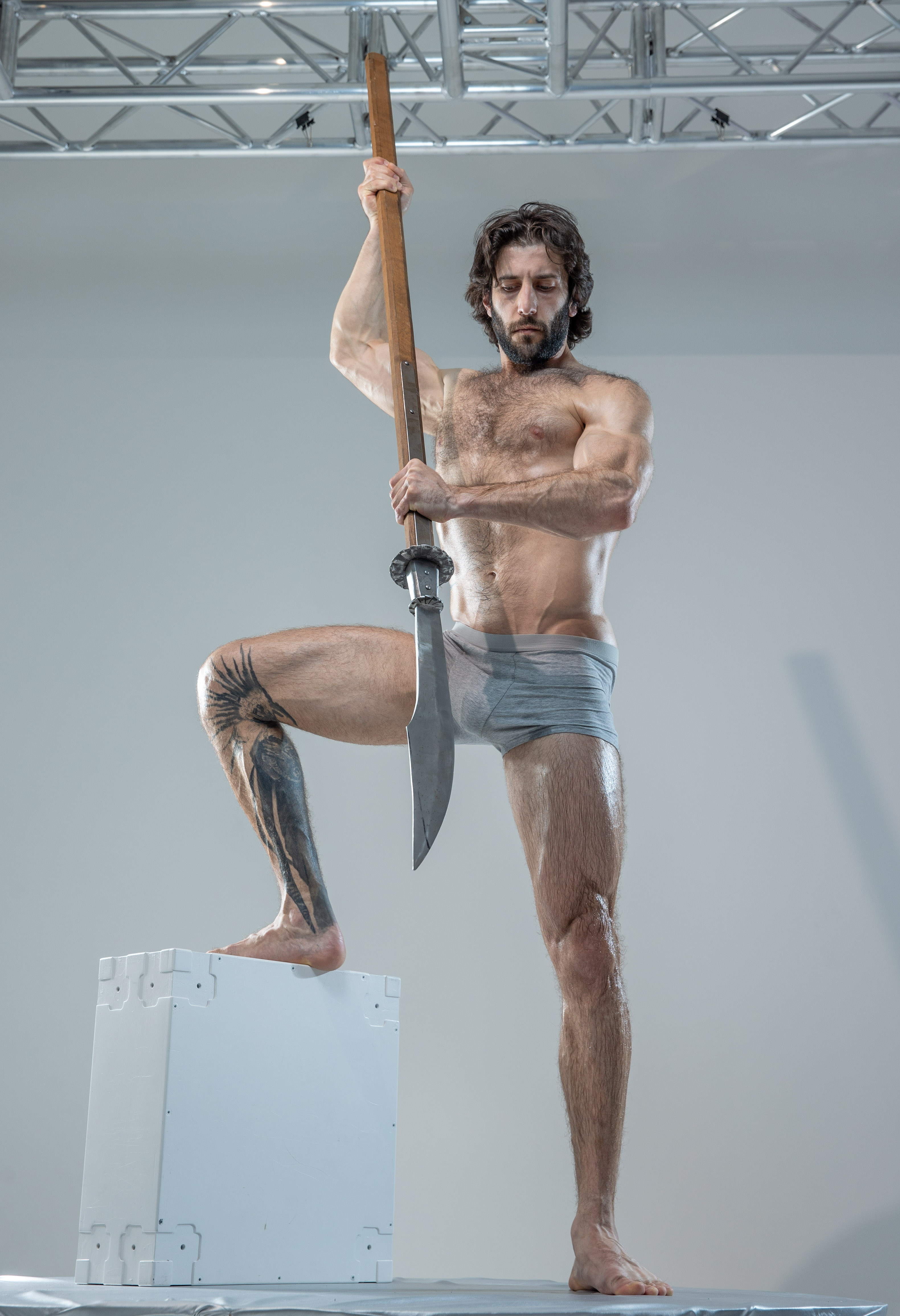 a muscular man with long, dark hair and beard stands on a white platform in a bright studio. he holds a long spear with a decorative blade aloft in a powerful pose. he's wearing only grey underwear, showcasing his physique and tattoos. the background is a neutral grey, focusing attention on the man and his spear.