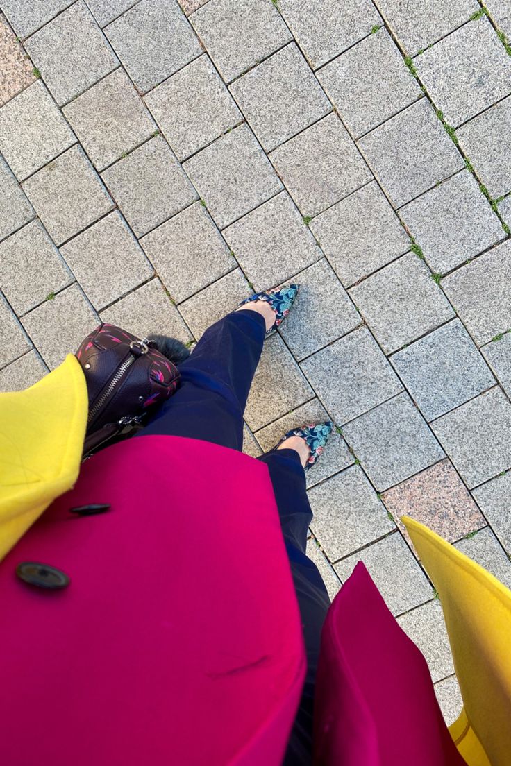 a highangle, closeup shot captures a woman's stylish outfit. she's wearing a vibrant bicolor coat, predominantly magenta with yellow accents. her navy trousers and floralprinted heels complement the coat's bold hues. a purple handbag with a decorative element rests partially visible beside her. the scene is set on a paved walkway, creating a striking contrast between the woman's bright clothing and the neutral gray of the stone tiles. the composition draws attention to both the vibrant fashion choices and the textured pavement.