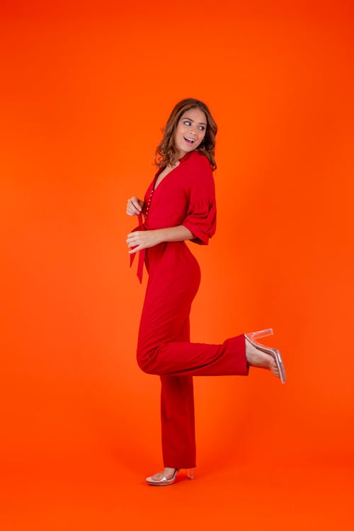 a woman in a red jumpsuit and clear heels is posing on an orange background. she has one leg raised and is looking back while smiling. the jumpsuit has a tie at the waist.