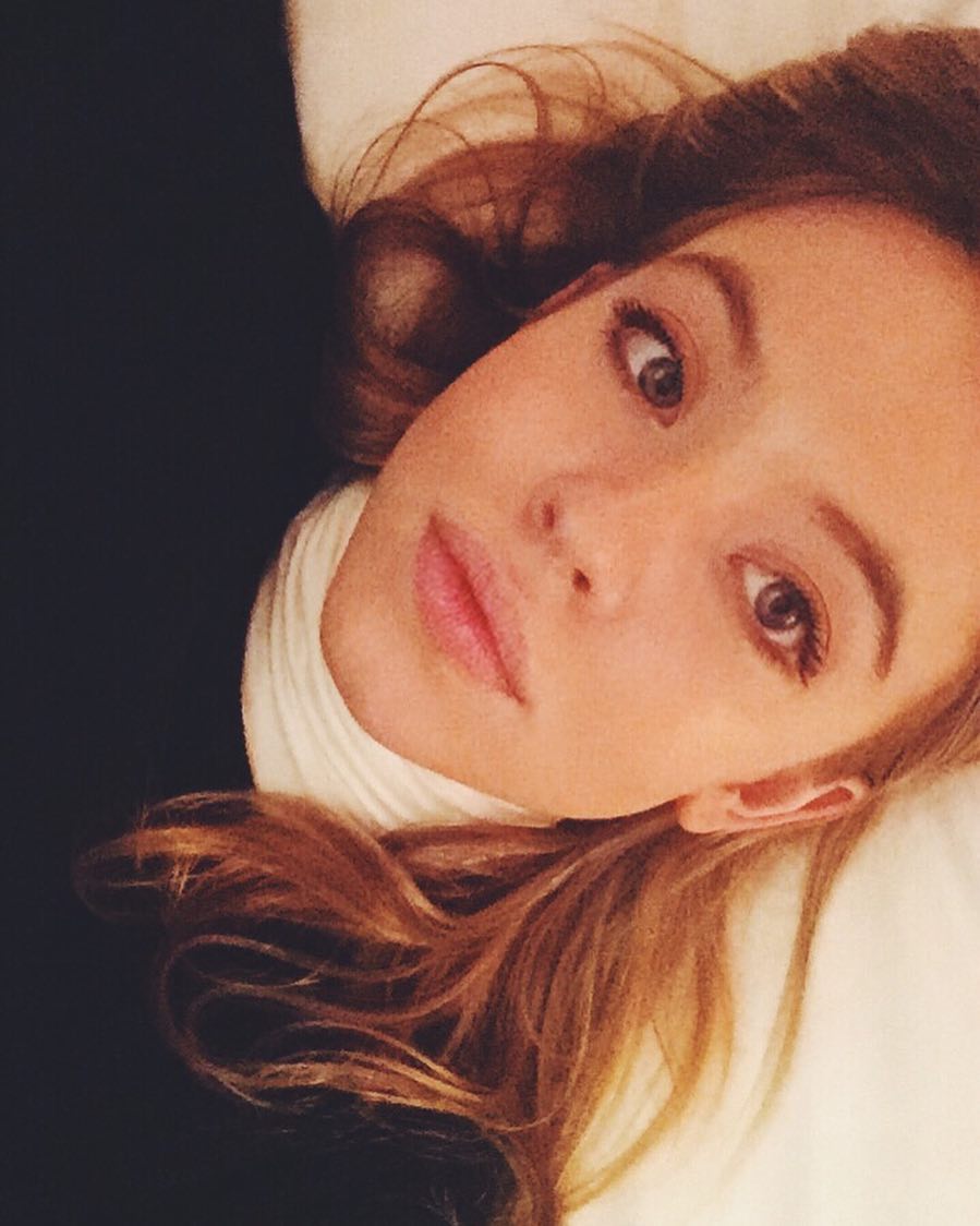 a closeup selfie of a young woman with long, light brown hair lying on her side on a bed.  she's wearing a black sweater and a white turtleneck. she's looking up at the camera, and her expression is serene and relaxed. the lighting is soft, and her makeup is minimal and natural. the overall mood of the image is calm and intimate.