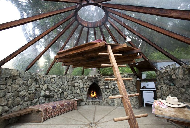 this is a shot of an interior space with a domeshaped roof made of wooden beams and clear panels. there is a stone wall surrounding the lower portion of the room, and a fireplace is visible with a fire burning inside. a loft bed is accessible by a wooden ladder. there is a bench covered with a carpet, a hat resting on a surface, and a small oven near a window. plants can be seen through the roof, providing natural light to the room. a geometric pattern is visible on the floor.
